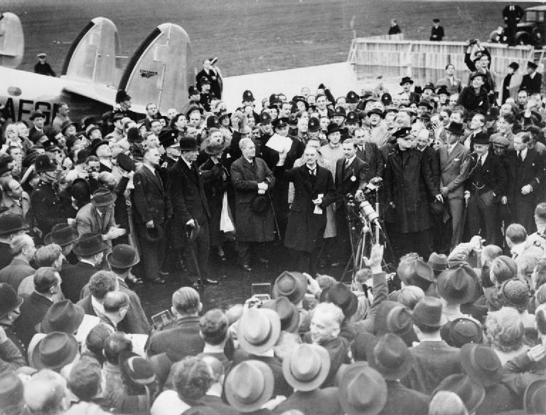 Photograph of Neville Chamberlain waving the Munich Agreement in his hand while standing in front of an airplane. 